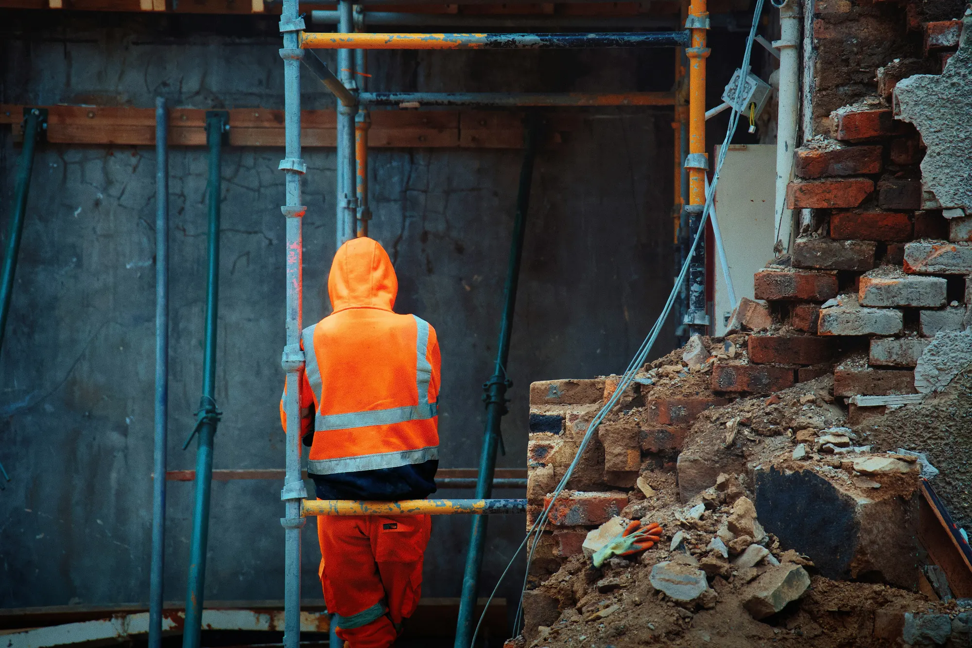 A worker seen from behind in a hooded orange hi-vis jacket and trousers, standing at a scaffold frame on a demolition site among brick rubble