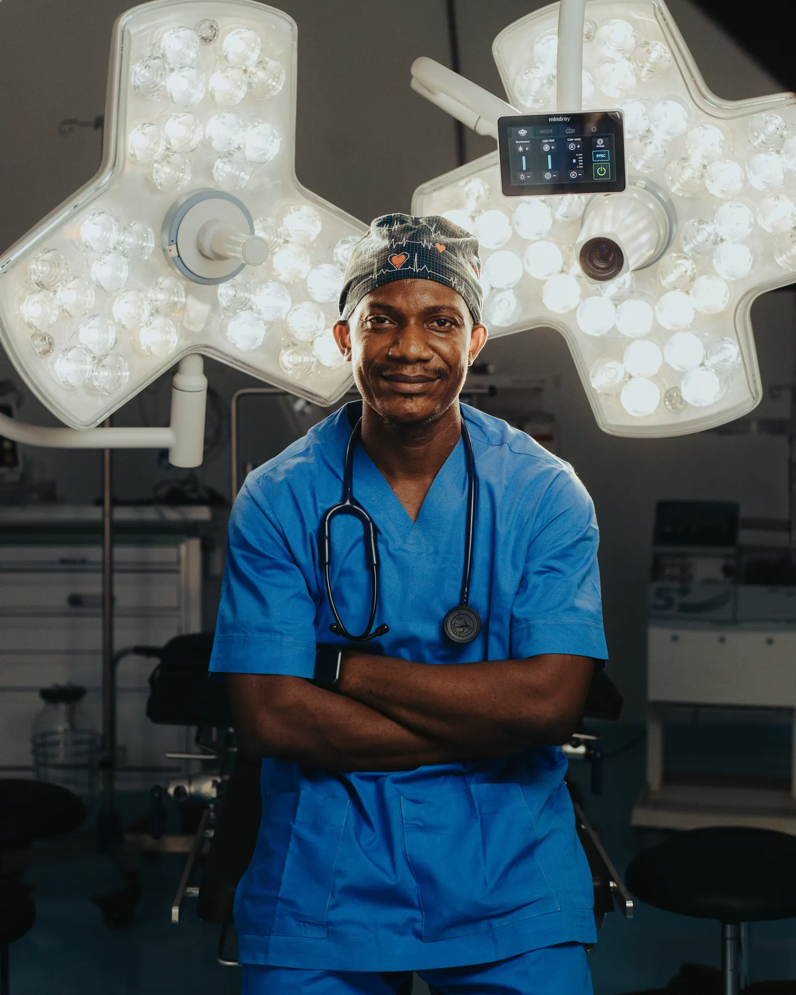 A clinician in blue scrubs and surgical cap with a stethoscope, standing under operating theatre lights