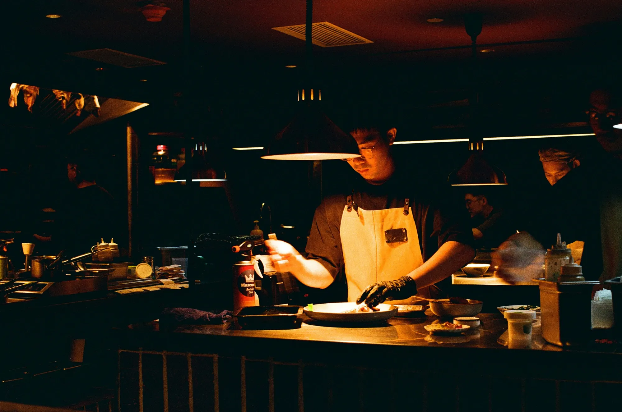 A cook in a dark tee and a heavy canvas apron plating under the heat lamps of an open kitchen pass