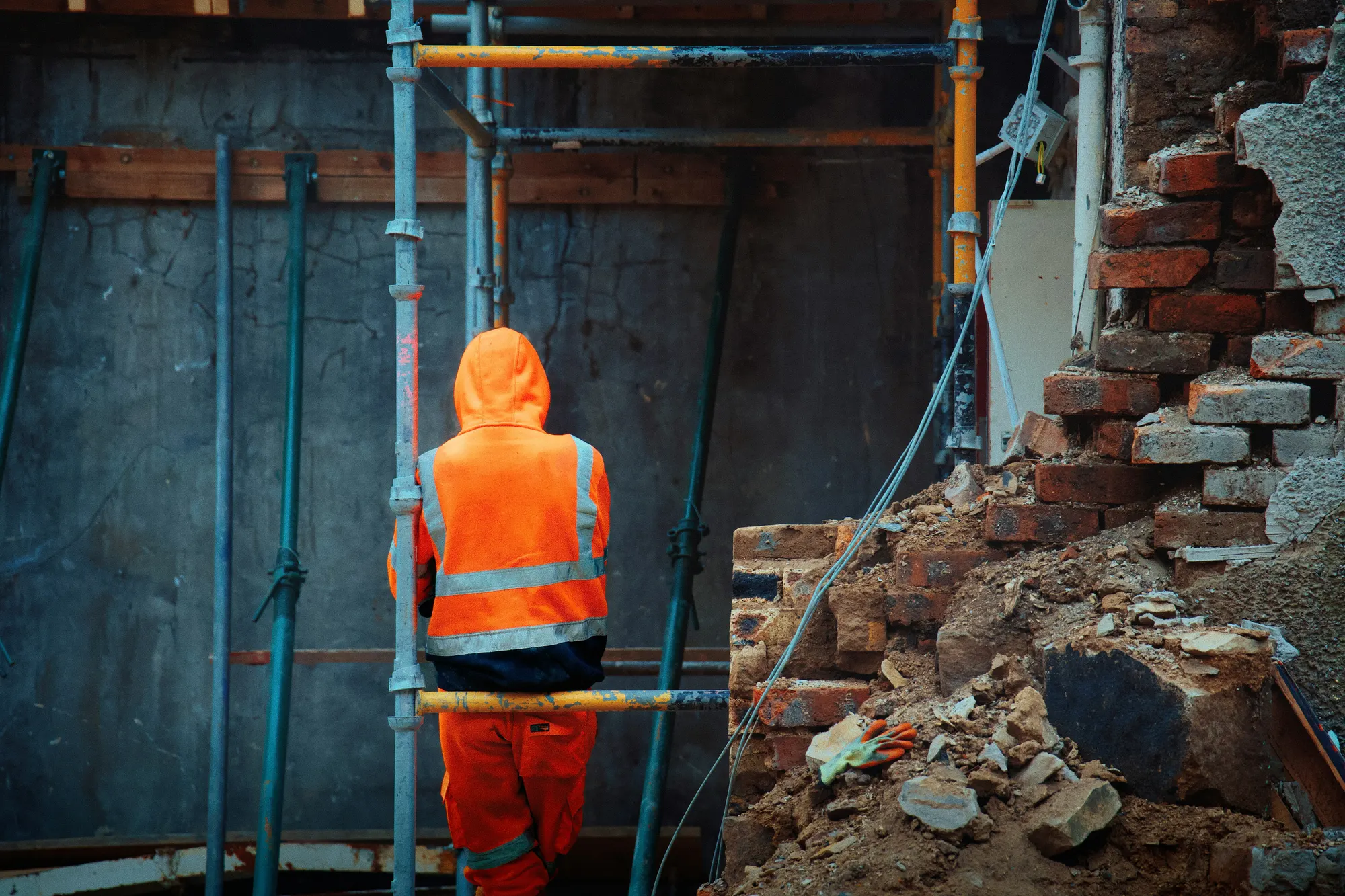 A worker seen from behind in a hooded orange hi-vis jacket and trousers, standing at a scaffold frame on a demolition site among brick rubble