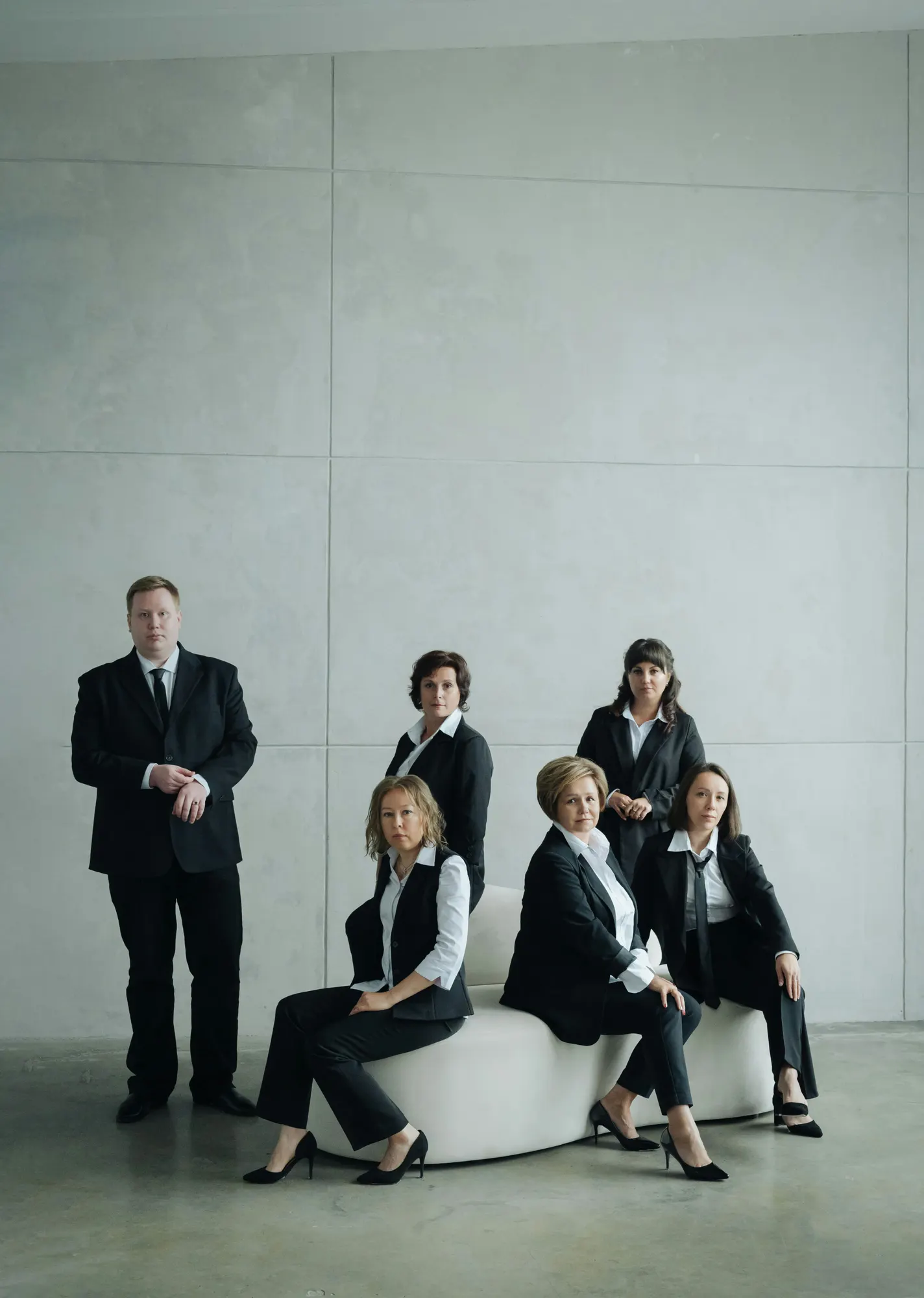 A line of staff in matching black suiting and waistcoats over white shirts, photographed straight on against a concrete wall